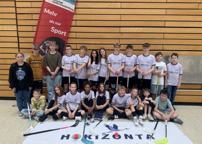 Alle Schüler:innen der AG Floorball der HORIZONTA mit Floorballschlägern vor einem Banner mit dem Logo der Schule. Die für das Turnier aufgestellten Schüler:innen in rosafarbenen Trikots.
