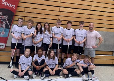 Die Floorball-Mannschaft der HORIZONTA in rosafarbenen Trikots mit Floorballschlägern vor einem Banner mit dem Logo der Schule mit ihrem betreuenden Lehrer.