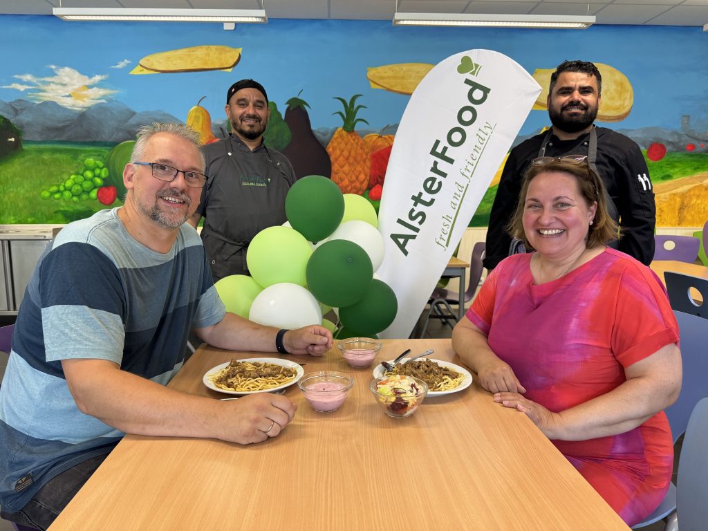 Schulleiterin Iris Strunk und Ganztagsbeauftragter Ulf Gerkensmeyer sitzen mit Essen an einem Tisch in der Mensa. Dahinter stehen zwei Mitarbeiter des neuen Mensabetreibers AlsterFood, in ihrer Mitte eine Fahne mit dem Logo der Firma und dekorative Ballons in den grünen Firmenfarben.