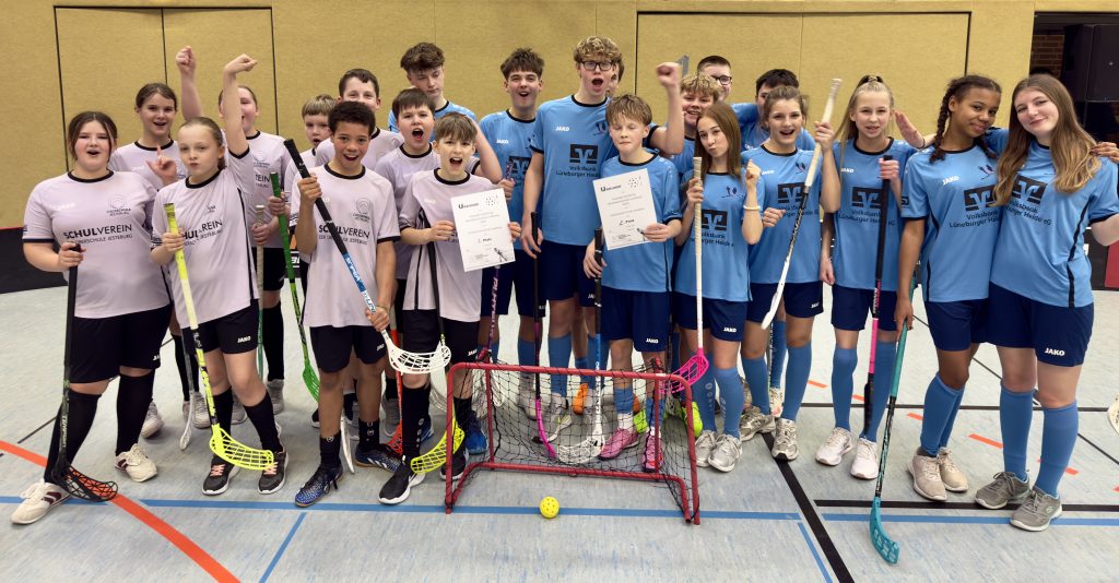 Unsere U14- und U16-Mannschaften mit Floorball-Schlägern und ihren Urkunden zum jeweils 2. Platz in ihren jeweiligen Trikots (rosa und blau) vor einem Floorball-Tor.